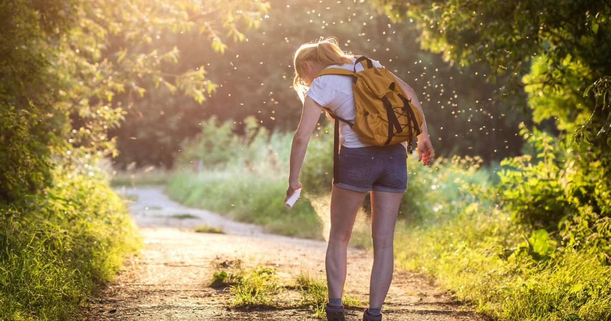 Eine Frau in der Natur, umgeben von Mücken. Mit einem Spray in der Hand.