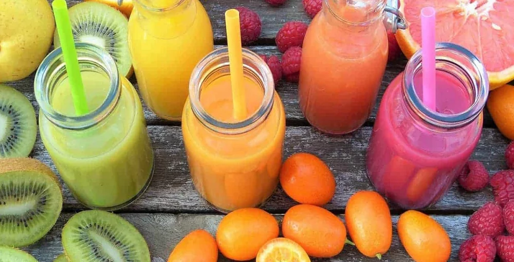 Orangefarbener, grüner und rosa Smoothie, umgeben von Früchten auf einem Holztisch