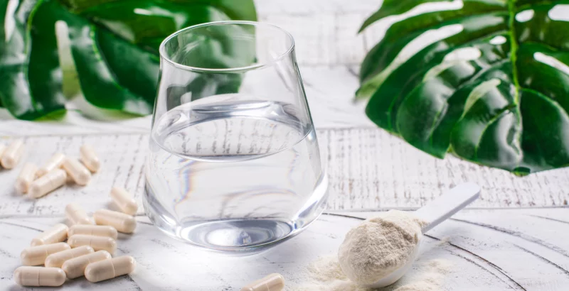Ein Glas Wasser mit Kapseln und Kollagen-Pulver daneben.
